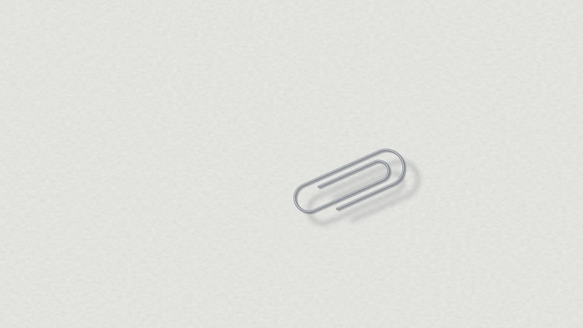 Paperclip: A single steel paperclip lies at an angle on a sheet of cool white paper, its long low shadow doing most of the drawing. Office stationery as a quiet still life.